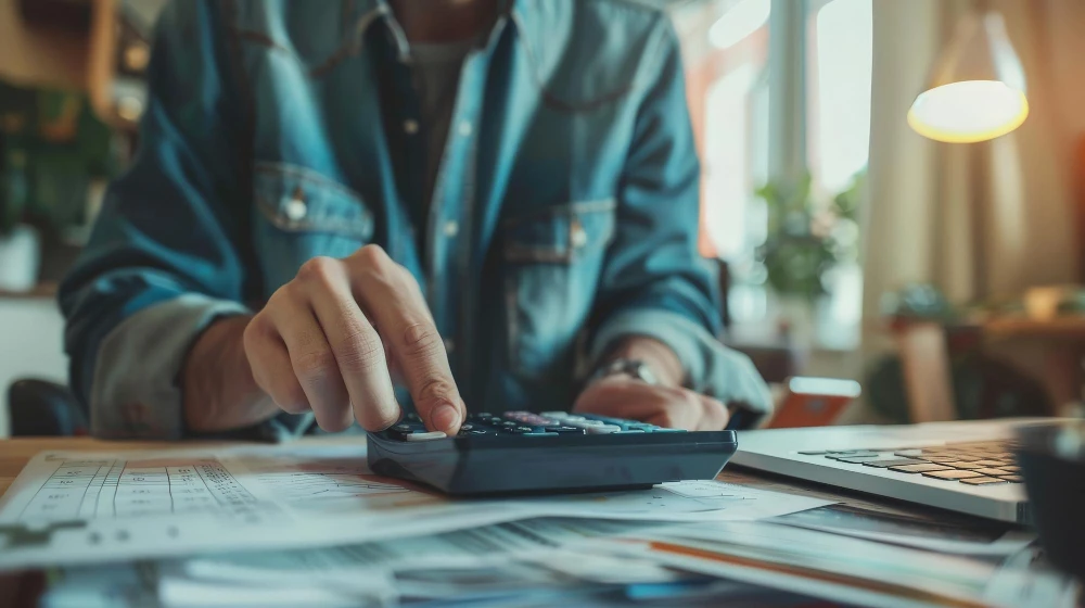 Caminhoneiro autônomo utilizando calculadora para fazer seu imposto de renda, com laptop e documentos ao lado, demonstrando independência e tecnologia