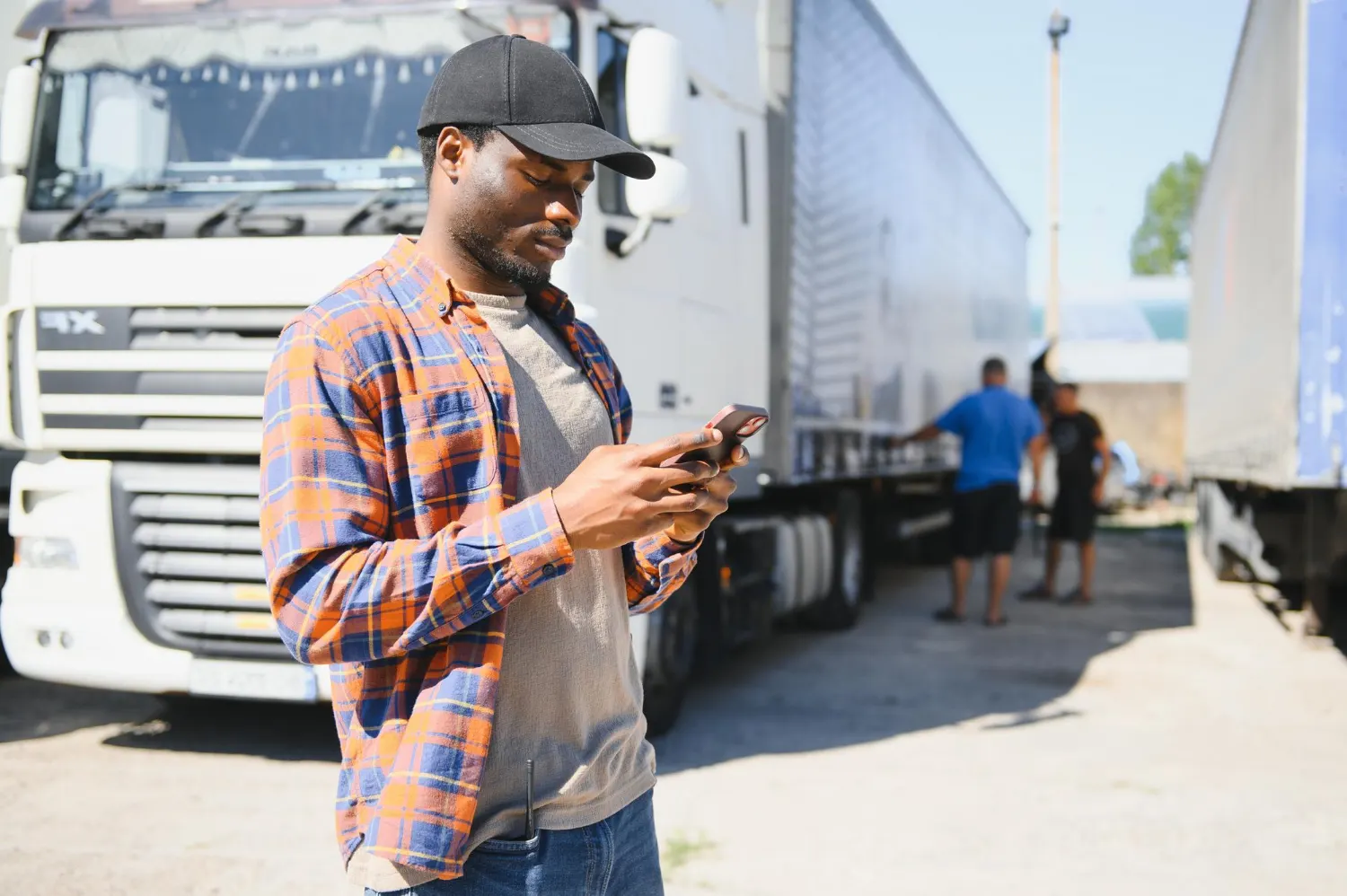 novas regras cnh: Trabalhador em pé ao lado de um caminhão, usando o celular em uma área externa de transporte e logística, sob luz solar.